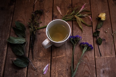 白色陶瓷杯，在绿叶植物和花瓣花旁边的木质表面上有茶
